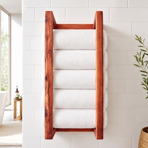 Op de afbeelding: Een houten handdoekenrek met een ladderontwerp, gebeitst in een warme roodbruine tint. Het rek bevat zes opgerolde witte handdoeken. Het rek is gemonteerd op een witte betegelde muur, wat een spa-achtige sfeer creëert. Ongeveer 60 cm breed.