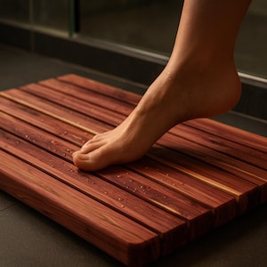May include: A rectangular bath mat constructed from parallel reddish-brown wooden slats. The surface of the mat features visible wood grain patterns and small water droplets scattered across the planks, providing a textured, slatted design.