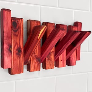 May include: A wall-mounted wooden coat rack with a rich red stain. The rack features multiple angled pegs for hanging items. The wood grain is visible, adding a natural aesthetic. The rack is mounted on a white tiled wall.