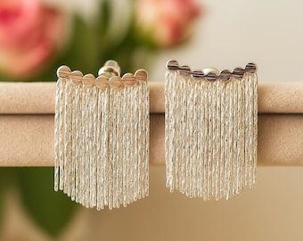 May include: A pair of silver-colored fringe earrings with a gold-colored scalloped top. The earrings are displayed on a neutral-colored surface, with blurred pink roses in the background. The earrings have a post backing.