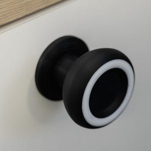 May include: A black cabinet knob with a white accent ring. The knob is round and has a flat base. The knob is mounted on a white surface.