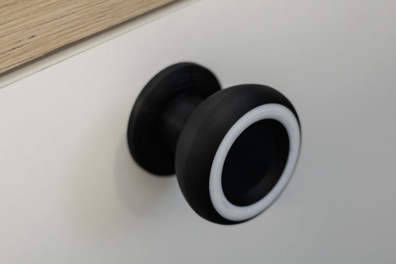 May include: A black cabinet knob with a white accent ring. The knob is round and has a flat base. The knob is mounted on a white surface.