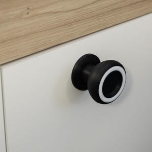 May include: A black cabinet knob with a white accent ring. The knob is mounted on a white cabinet door. The top of the cabinet is light brown wood.