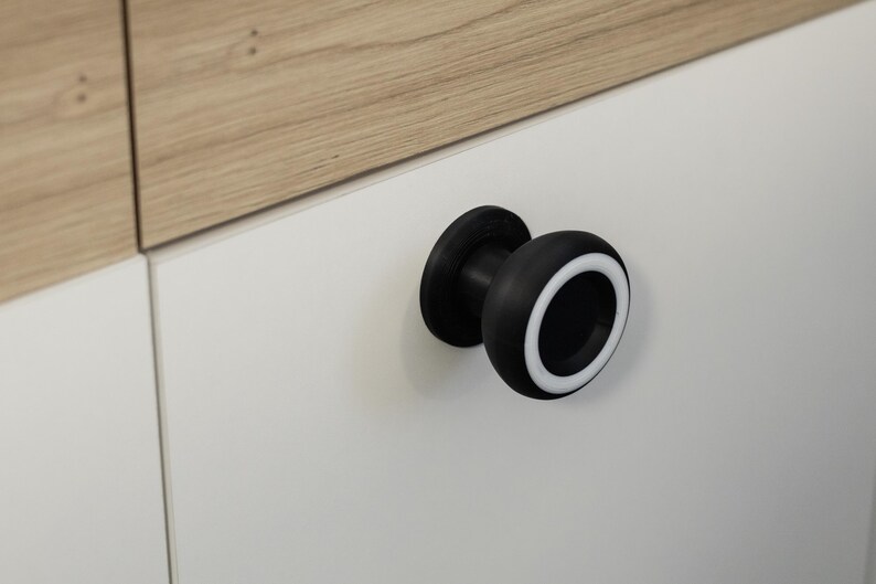 May include: A black cabinet knob with a white accent ring. The knob is mounted on a white cabinet door. The top of the cabinet is light brown wood.