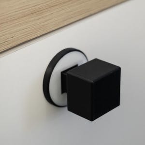 May include: A black cube cabinet knob with a white and black circular base. The knob is mounted on a white surface, with a section of light wood visible in the upper left corner.