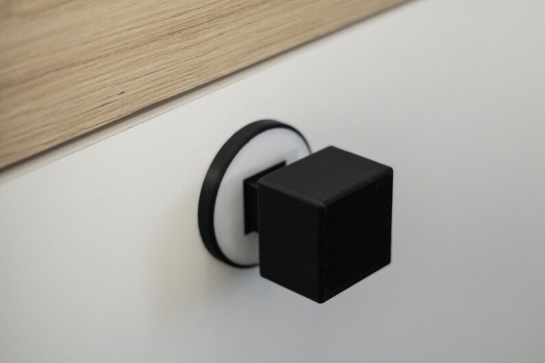 May include: A black cube cabinet knob with a white and black circular base. The knob is mounted on a white surface, with a section of light wood visible in the upper left corner.