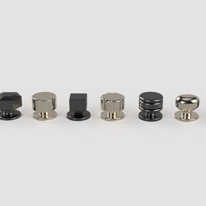 May include: A selection of ten cabinet knobs in diverse shapes and finishes. The knobs feature round, faceted, and cylindrical designs in metallic colours such as black, silver, and gold. They are arranged in a row against a white backdrop.