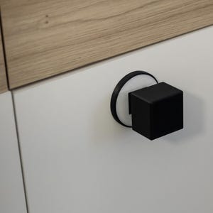 May include: A close-up of a modern cabinet door with a black cube-shaped handle. The handle is mounted on a white circular base. The cabinet door is white, and the top panel is light brown wood.