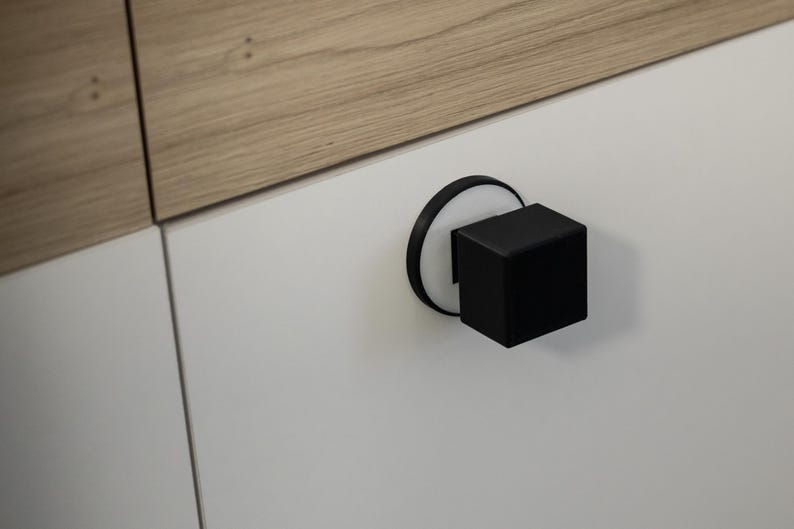 May include: A close-up of a modern cabinet door with a black cube-shaped handle. The handle is mounted on a white circular base. The cabinet door is white, and the top panel is light brown wood.
