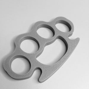 May include: A gray, cast-metal knuckle duster with four finger holes and a curved grip. The weapon is shown against a white background. The knuckles are a self-defense tool.