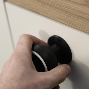 May include: A black and white cabinet knob with a circular design. The knob is held by a hand and is attached to a white cabinet door. The top of the cabinet is wood.