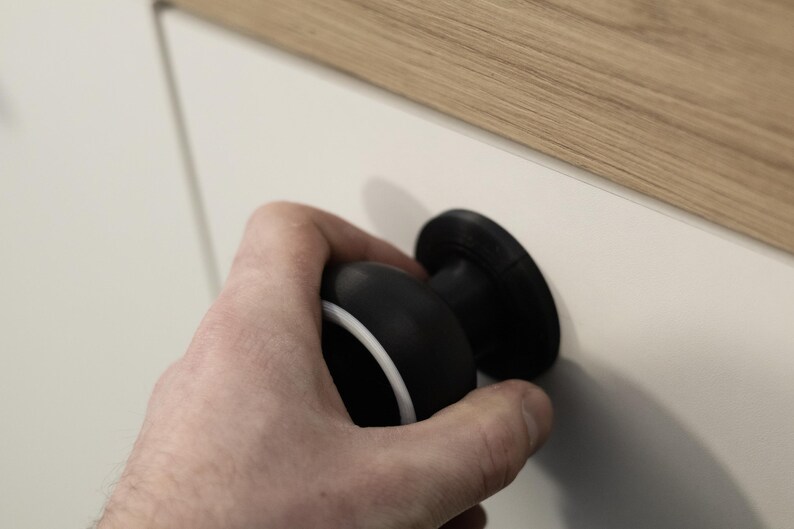 May include: A black and white cabinet knob with a circular design. The knob is held by a hand and is attached to a white cabinet door. The top of the cabinet is wood.
