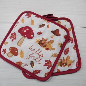 Könnte beinhalten: Zwei quadratische Topflappen mit weißem Hintergrund, mit rot gekappten Pilzen, Herbstblättern und dem Text "hello fall". Die Topflappen haben einen roten Rand.