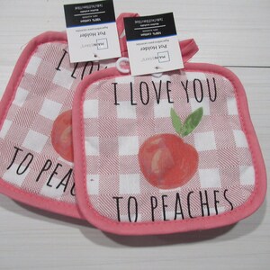 May include: Two pink and white gingham pot holders with the text "I LOVE YOU TO PEACHES" and a peach illustration. The pot holders have a pink border and a loop for hanging. Tags are attached.