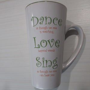 May include: White ceramic latte mug with a curved handle. The mug features the words "Dance", "Love", and "Sing" in green script, with additional text in red. The mug is approximately 6 inches tall.