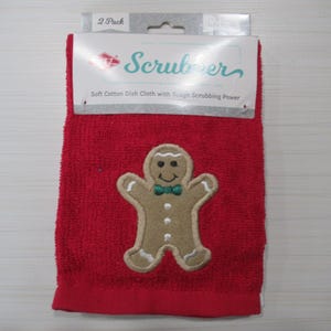 May include: Red kitchen towel with an embroidered gingerbread man design. The towel has the word "Scrubber" printed on the top. The gingerbread man has a green bow tie and white accents.