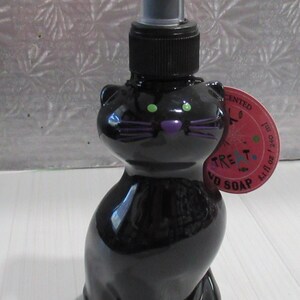 Op de afbeelding: Zwarte katvormige handzeepdispenser met een pomptop. De kat heeft groene ogen en paarse snorharen en een neus. Een rond label is aan de fles bevestigd. De fles staat op een wit oppervlak.