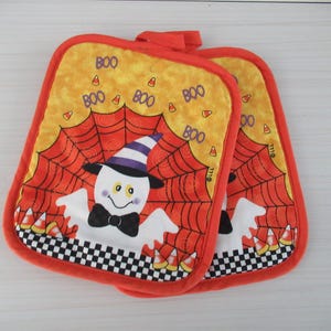 Puede incluir: Dos agarraderas con temática de Halloween. Cada una presenta un fantasma sonriente con sombrero de bruja y pajarita, sobre un diseño de telaraña. El fondo es naranja y amarillo con la palabra "BOO" impresa. Los bordes son rojos.