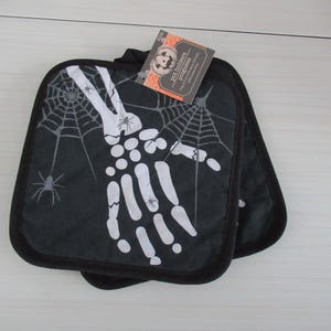 Puede incluir: Dos agarraderas negras con un diseño de mano de esqueleto blanco y telaraña. Las agarraderas tienen un borde negro y una etiqueta que dice "pot holders" y "poignées". Accesorios de cocina con temática de Halloween.