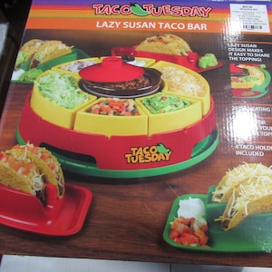 Puede incluir: Un juego de barra de tacos Lazy Susan rojo, amarillo y verde. El juego incluye una olla de calentamiento central, compartimentos para ingredientes y soportes para tacos. La caja dice "TACO TUESDAY LAZY SUSAN TACO BAR".