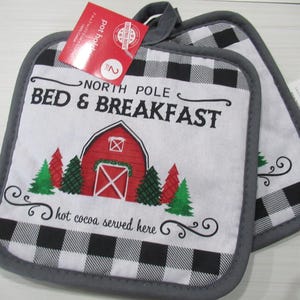 May include: Two square pot holders with a black and white buffalo check pattern border and a gray trim. The center features a red barn illustration with green and red trees, and the text "NORTH POLE BED & BREAKFAST" and "hot cocoa served here."