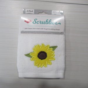 Puede incluir: Paño de cocina de algodón blanco con una aplicación de girasol amarillo y hojas verdes. El paño de cocina tiene la palabra "Scrubber" impresa en el embalaje. El embalaje indica que es un paquete de 2.