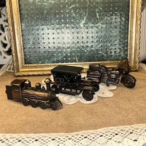 May include: Three bronze-colored miniature vehicles: a train, a vintage car, and a motorcycle. The train is on the left, the car in the middle, and the motorcycle on the right. They are placed on a burlap surface with a framed picture in the background.