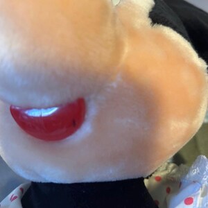 May include: Close-up of a plush toy featuring a character with a light pink face, red lips, and a black collar. The toy has a white bow with red polka dots and a white dress with red polka dots.