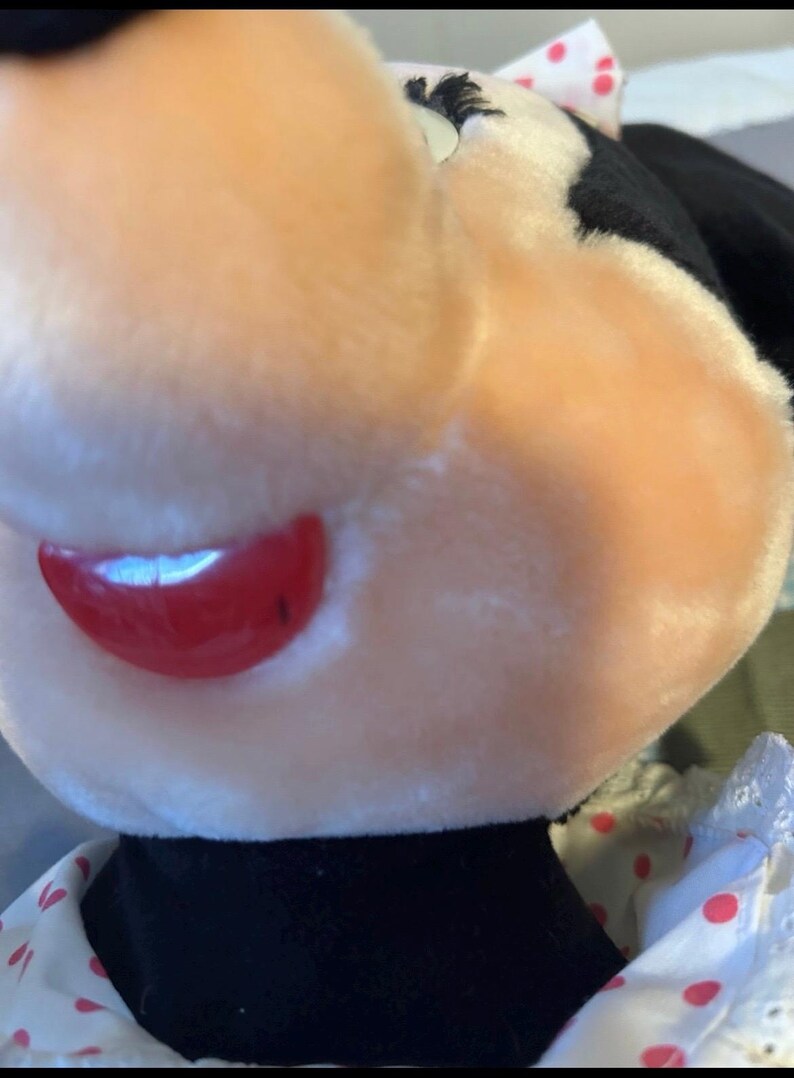 May include: Close-up of a plush toy featuring a character with a light pink face, red lips, and a black collar. The toy has a white bow with red polka dots and a white dress with red polka dots.
