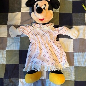 May include: A plush Minnie Mouse doll wearing a pink polka dot dress with a white lace trim. The doll has black ears, a pink bow, and yellow shoes. The doll is lying on a patchwork quilt.