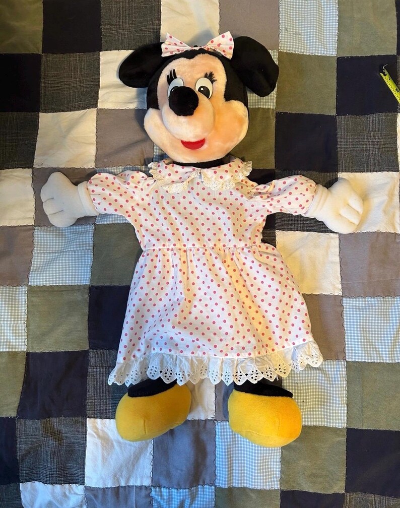 May include: A plush Minnie Mouse doll wearing a pink polka dot dress with a white lace trim. The doll has black ears, a pink bow, and yellow shoes. The doll is lying on a patchwork quilt.