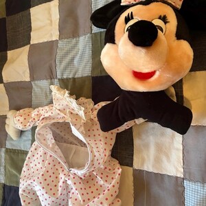 May include: A plush Minnie Mouse head with a pink bow and a matching polka dot outfit. The outfit is white with red polka dots and lace trim. The plush toy has black ears and a red mouth.
