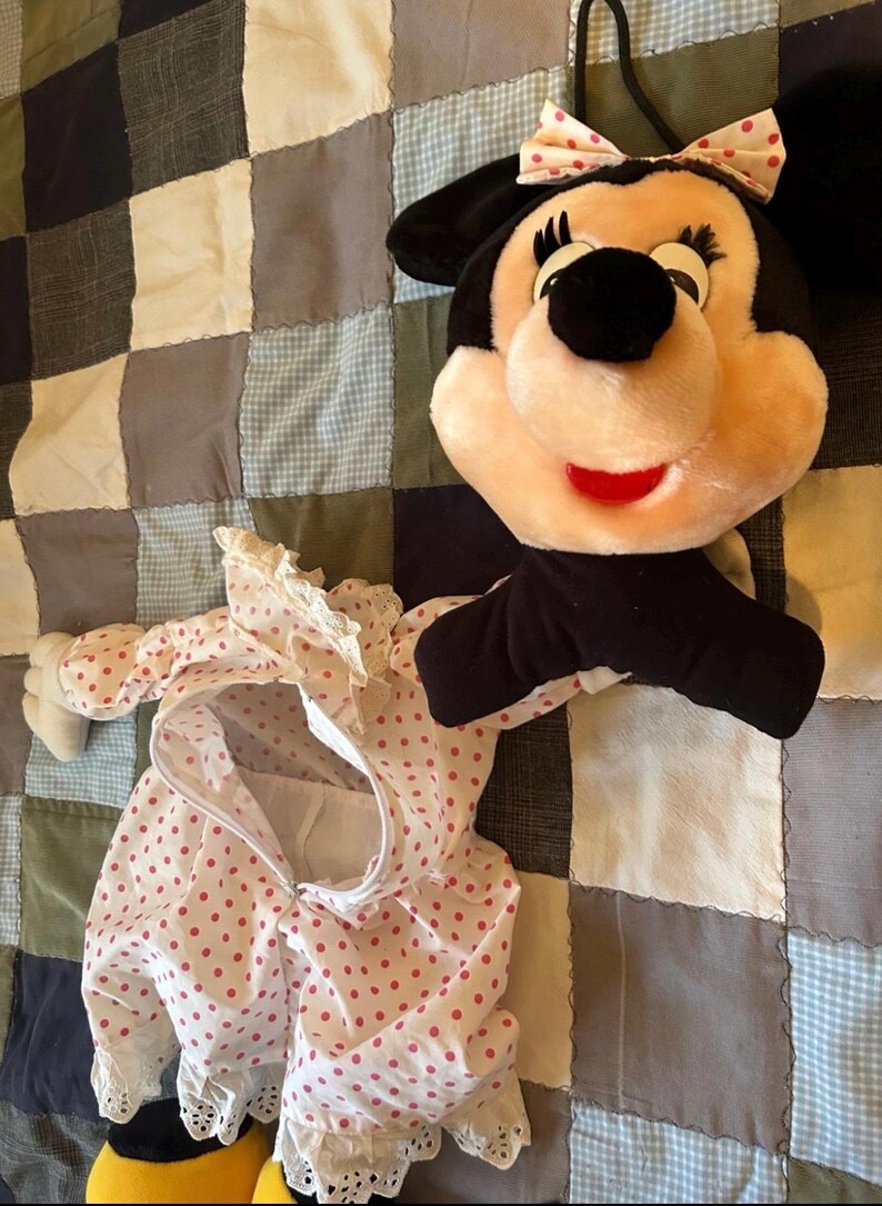 May include: A plush Minnie Mouse head with a pink bow and a matching polka dot outfit. The outfit is white with red polka dots and lace trim. The plush toy has black ears and a red mouth.