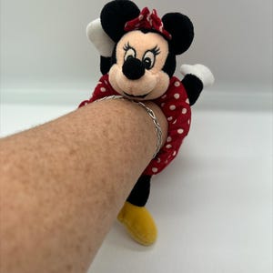 Puede incluir: Un peluche de Minnie Mouse unido a una goma de pelo de tela roja con lunares blancos, con un lazo rojo de lunares blancos, orejas negras, guantes blancos y zapatos amarillos, llevado con una fina pulsera de cadena de metal plateado.