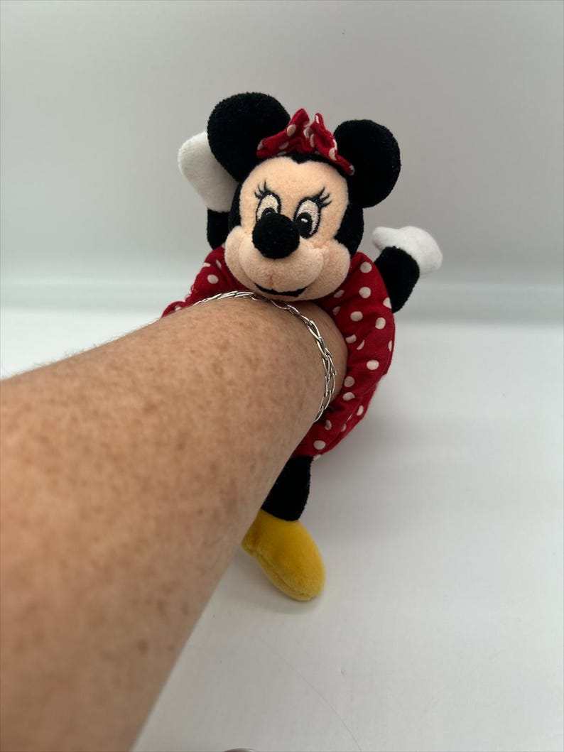 Puede incluir: Un peluche de Minnie Mouse unido a una goma de pelo de tela roja con lunares blancos, con un lazo rojo de lunares blancos, orejas negras, guantes blancos y zapatos amarillos, llevado con una fina pulsera de cadena de metal plateado.