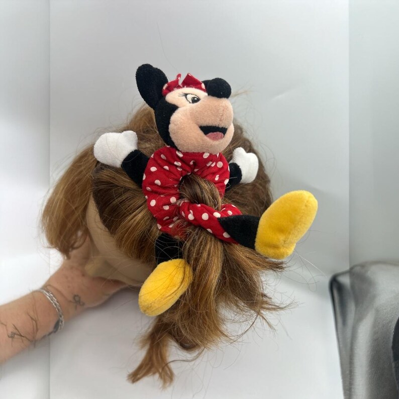 Puede incluir: Un coletero de felpa con un personaje de rat&oacute;n de dibujos animados con orejas negras, una banda de tela roja con lunares blancos, guantes blancos y zapatos amarillos. El personaje est&aacute; unido al coletero, que rodea un cabello casta&ntilde;o.