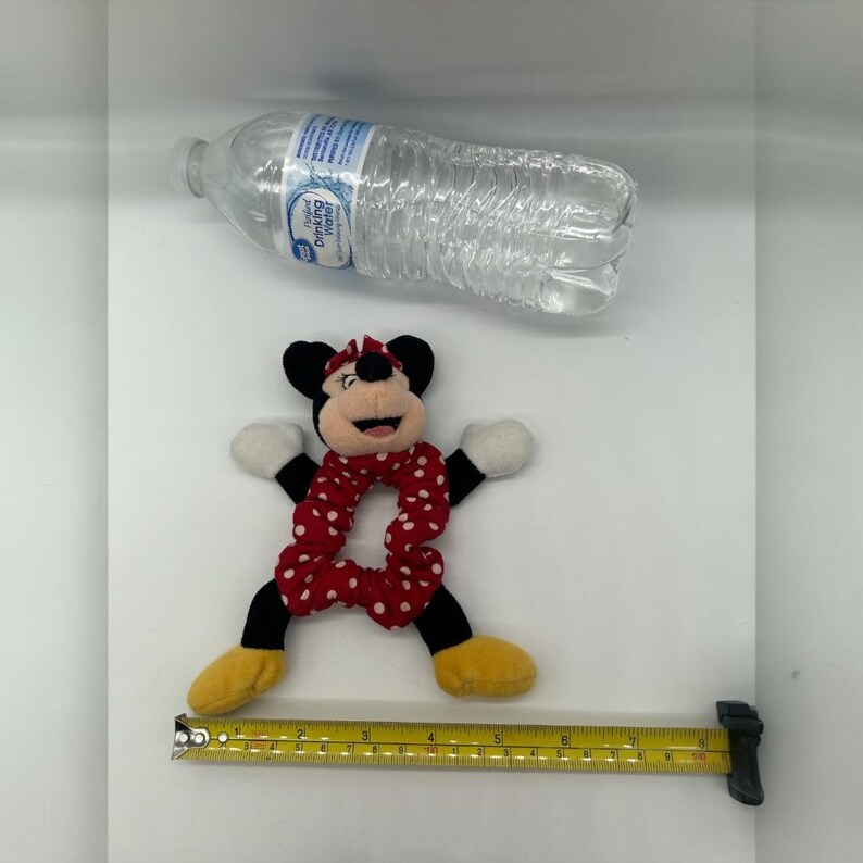 Puede incluir: Un coletero de peluche de Minnie Mouse con una banda de tela de lunares rojos y blancos, orejas negras, guantes blancos y zapatos amarillos. Se ve una botella de agua de pl&aacute;stico transparente y una cinta m&eacute;trica amarilla de 20 cm.