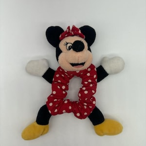 Puede incluir: Un coletero de peluche de Minnie Mouse con una banda de tela roja con lunares blancos. La cabeza del personaje tiene orejas negras, un lazo rojo y una cara color canela. Incluye brazos negros con guantes blancos y piernas negras con zapatos amarillos.