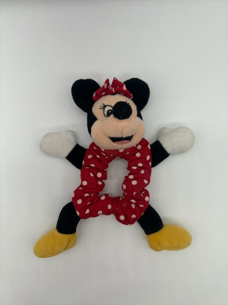 Puede incluir: Un coletero de peluche de Minnie Mouse con una banda de tela roja con lunares blancos. La cabeza del personaje tiene orejas negras, un lazo rojo y una cara color canela. Incluye brazos negros con guantes blancos y piernas negras con zapatos amarillos.