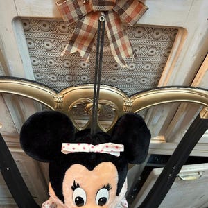 May include: A Minnie Mouse plush doll with large black ears and a pink bow with white polka dots. The doll is hanging from a decorative bow with brown and orange plaid fabric. The background includes a gold-colored ornate frame.