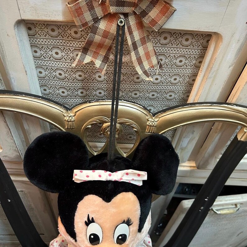 May include: A Minnie Mouse plush doll with large black ears and a pink bow with white polka dots. The doll is hanging from a decorative bow with brown and orange plaid fabric. The background includes a gold-colored ornate frame.