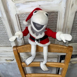 以下が含まれることがあります： サンタクロースの衣装を着た白いカエルのぬいぐるみ。赤いジャケットに白いトリム、黒いベルト、赤い帽子が含まれます。カエルは白い手袋と水かきのある足をしており、木製の椅子に座っています。