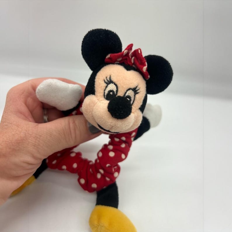 Puede incluir: Un coletero de felpa con la cabeza del personaje Minnie Mouse, orejas negras, un lazo rojo con lunares blancos y una banda de tela a juego con lunares blancos. El personaje tiene cara beige, nariz negra y manos con guantes blancos.