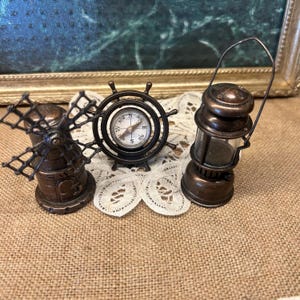 May include: A collection of miniature bronze-colored nautical-themed decorative items. Includes a windmill, a ship's wheel compass, and a lantern. The items are arranged on a white lace doily and a burlap surface, with a framed painting in the background.