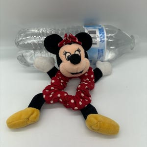 Puede incluir: Un peluche de Minnie Mouse unido a una goma de pelo de tela roja con lunares blancos. El personaje tiene orejas negras, un lazo rojo con lunares blancos, guantes blancos y zapatos amarillos. Detr&aacute;s se ve una botella de agua de pl&aacute;stico transparente.