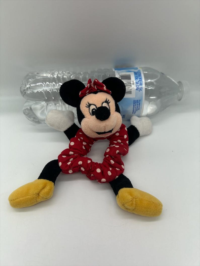 Puede incluir: Un peluche de Minnie Mouse unido a una goma de pelo de tela roja con lunares blancos. El personaje tiene orejas negras, un lazo rojo con lunares blancos, guantes blancos y zapatos amarillos. Detr&aacute;s se ve una botella de agua de pl&aacute;stico transparente.