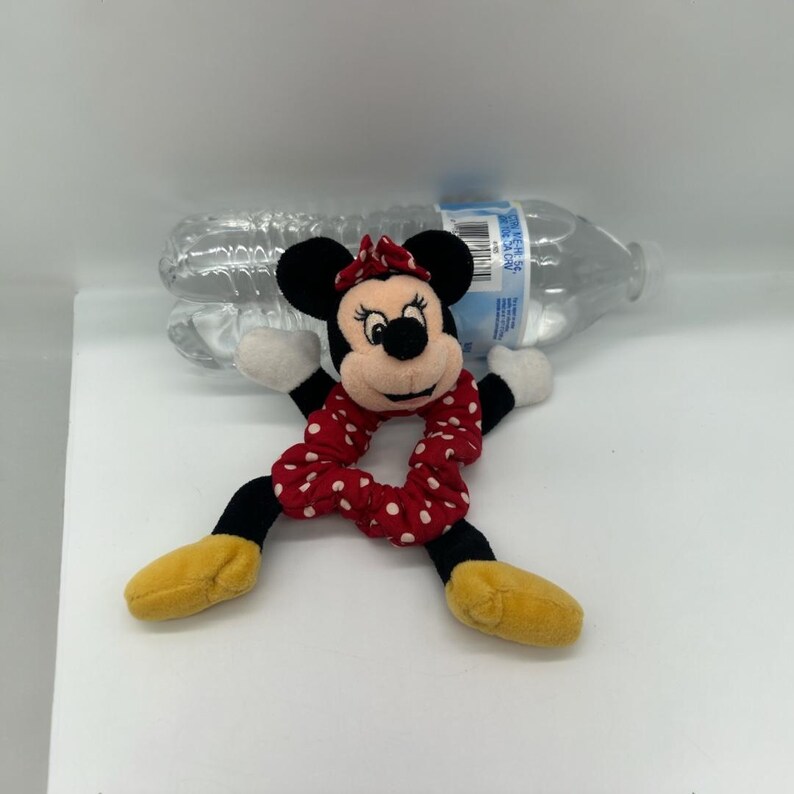 Puede incluir: Un coletero de peluche de Minnie Mouse con tela roja de lunares blancos, orejas negras, un lazo rojo, manos blancas y zapatos amarillos, colocado frente a una botella de agua de pl&aacute;stico transparente como referencia de escala.