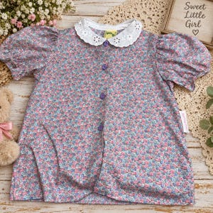 Op de afbeelding: Een blouse met korte mouwen voorzien van een blauw, roze en wit bloemenpatroon, een witte kanten kraag met oogjes en vier paarse knopen aan de voorzijde. Het kledingstuk heeft pofmouwen en een rechte zoom in een klassiek ontwerp.