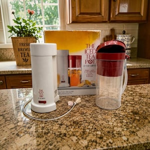 Puede incluir: Una máquina de té helado Mr. Coffee blanca con interruptor rojo, una jarra de plástico transparente con tapa roja y su caja de cartón original. La caja muestra el texto The Iced Tea Pot by Mr. Coffee e imágenes del aparato y té helado.