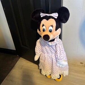 May include: A plush Minnie Mouse doll wearing a pink polka-dot dress with white lace trim and a matching bow. The doll has black ears, a peach-colored face, and yellow shoes. The doll is standing in front of a black door.